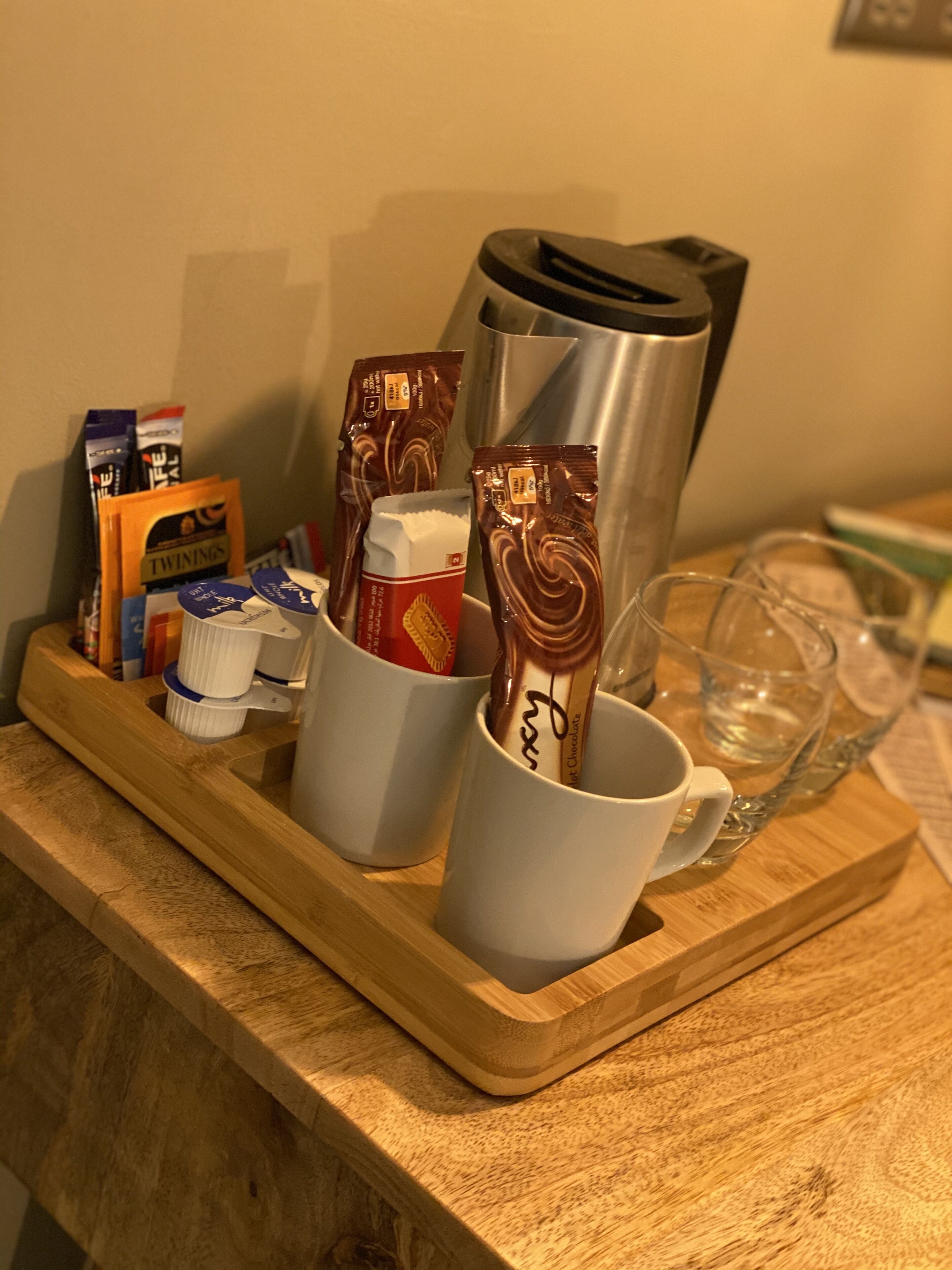 Ein Holztablett mit einer Kaffeemaschine, zwei Tassen, einem Glas und verschiedenen Kaffeekapseln auf einer Holz Arbeitsplatte.