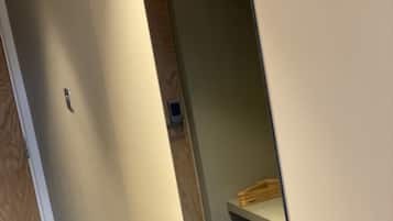 A narrow hallway with a wooden door on the left and a mirror reflecting a small bathroom area with a wooden shelf.