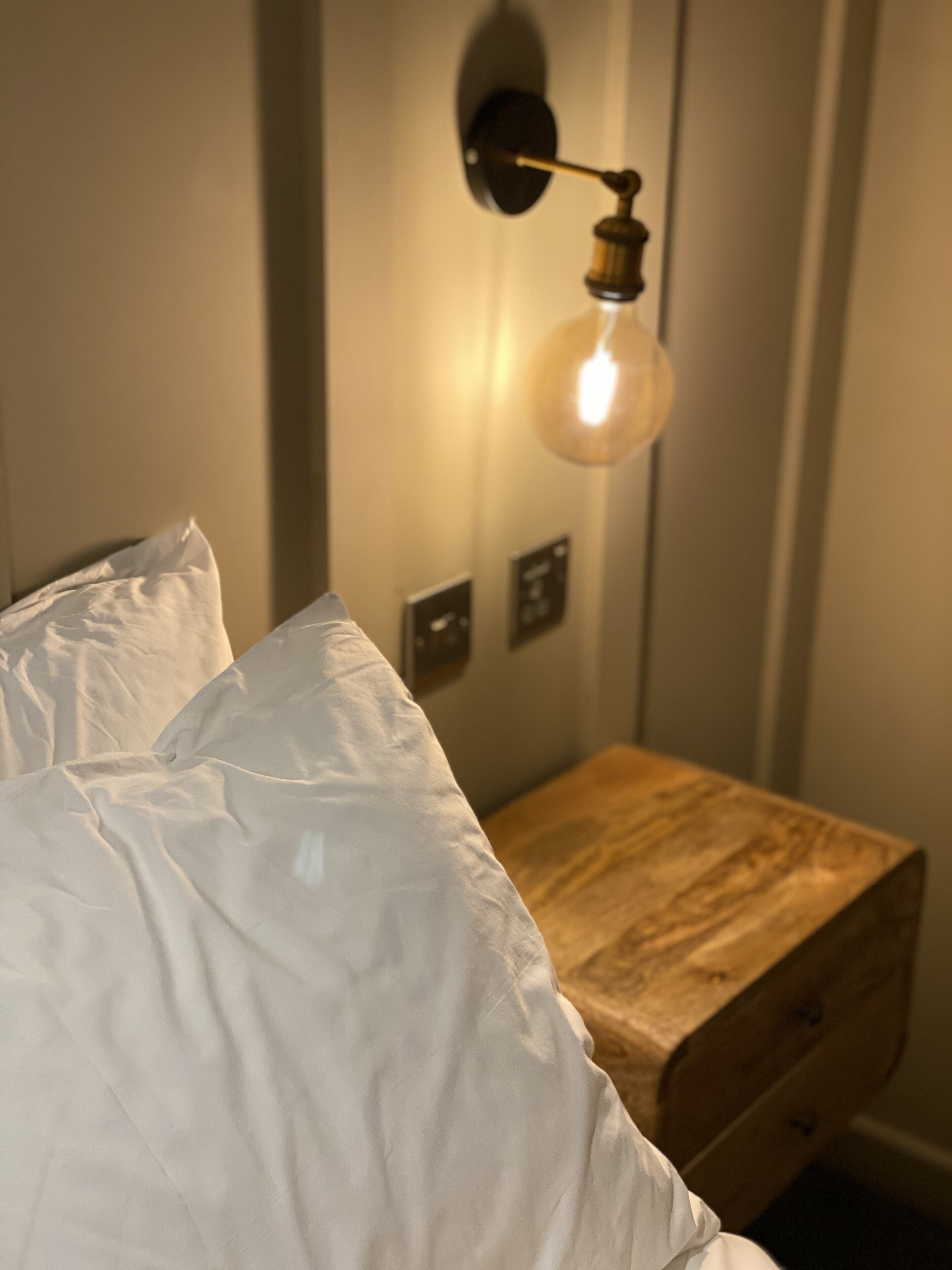 Ein Bett mit weißen Leinen, ein Nachttisch aus Holz mit Schubladen und eine an der Wand befestigte Leuchte.