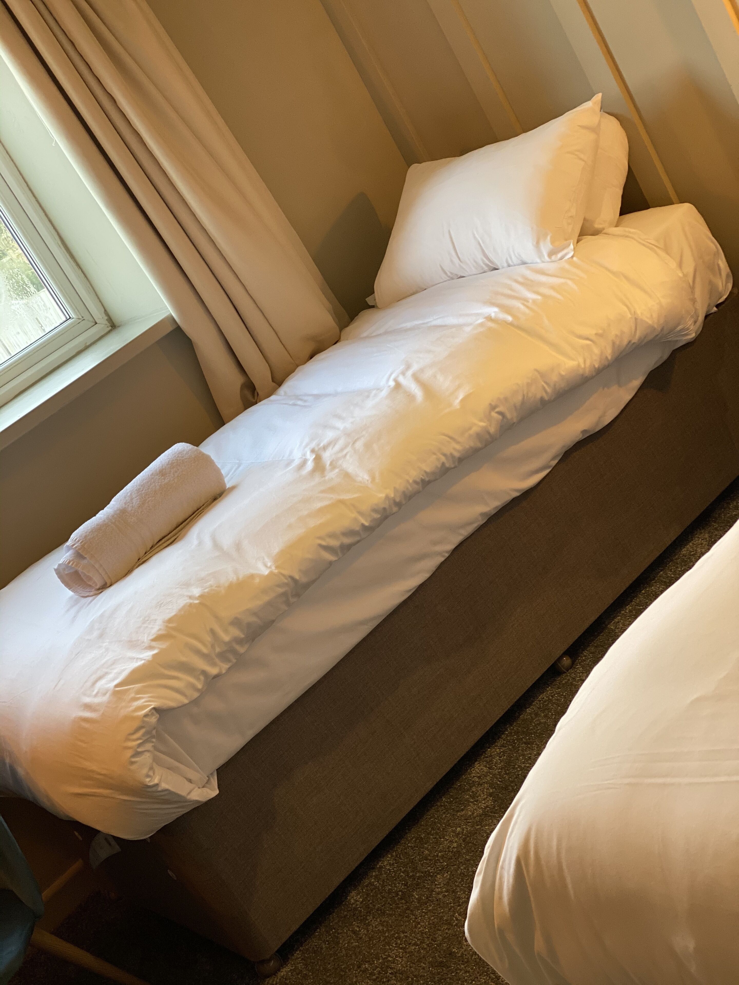 Ein Einzelbett mit weißer Bettwäsche und einem zusammengerollten Handtuch, neben einem mit Vorhängen versehenen Fenster.