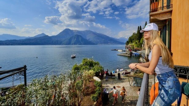A balcony overlooking a lake with boats, a mountain in the background, and people on the shore.