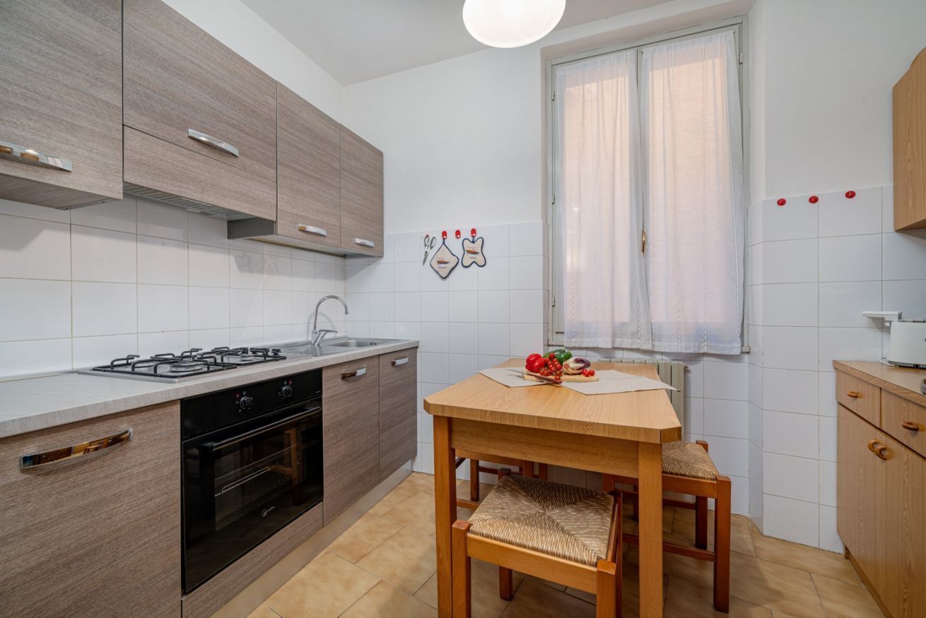 Una cucina moderna con piano cottura a gas, forno e mobili in legno. C'è un tavolo da pranzo con una sedia e una finestra con tende.
