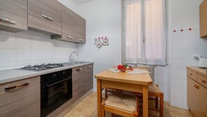 A modern kitchen with a gas stove, oven, and wooden cabinets. There is a dining table with a chair and a window with curtains.