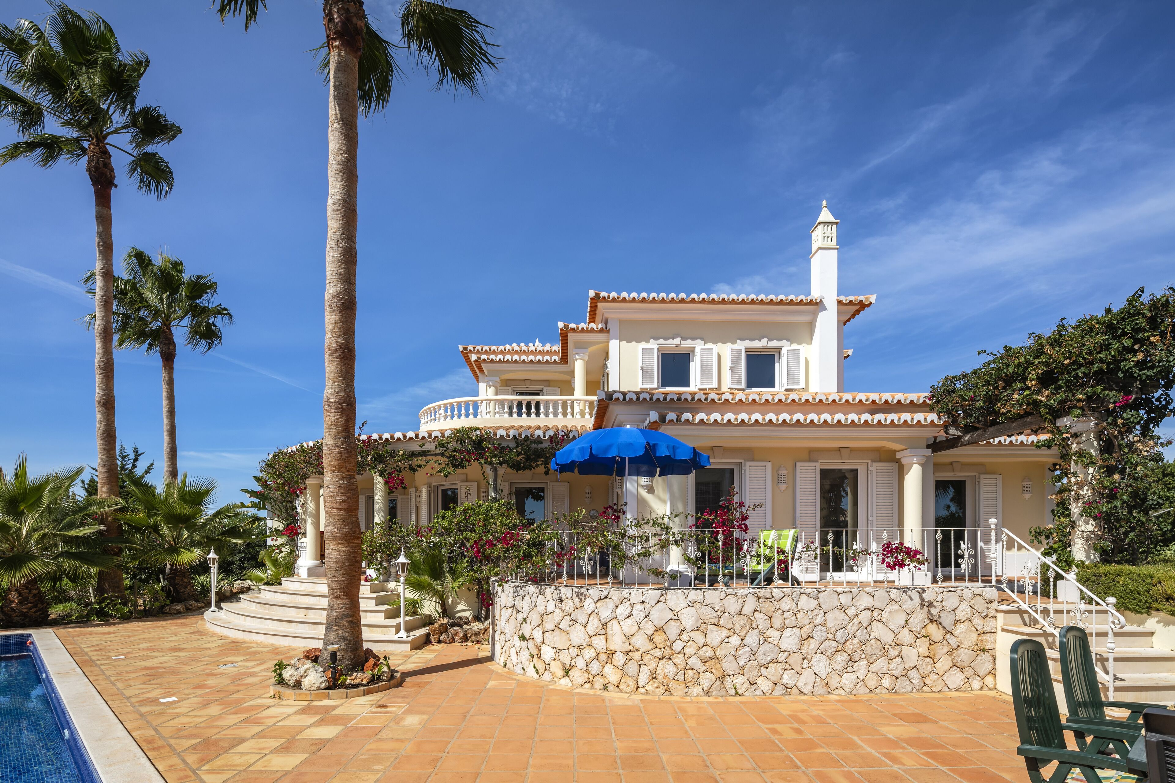 A two-story house with a stone wall, a pool, and palm trees.