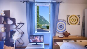 A room with a TV, a fan, a window with curtains, a wooden table, and wall art. - Casa Royal beautiful Garden (Varenna)