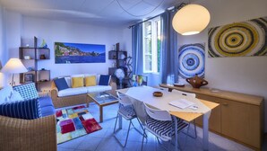 A modern living room with a sofa, a dining table, and a TV. There is a painting on the wall and a window with curtains. - Casa Royal beautiful Garden (Varenna)