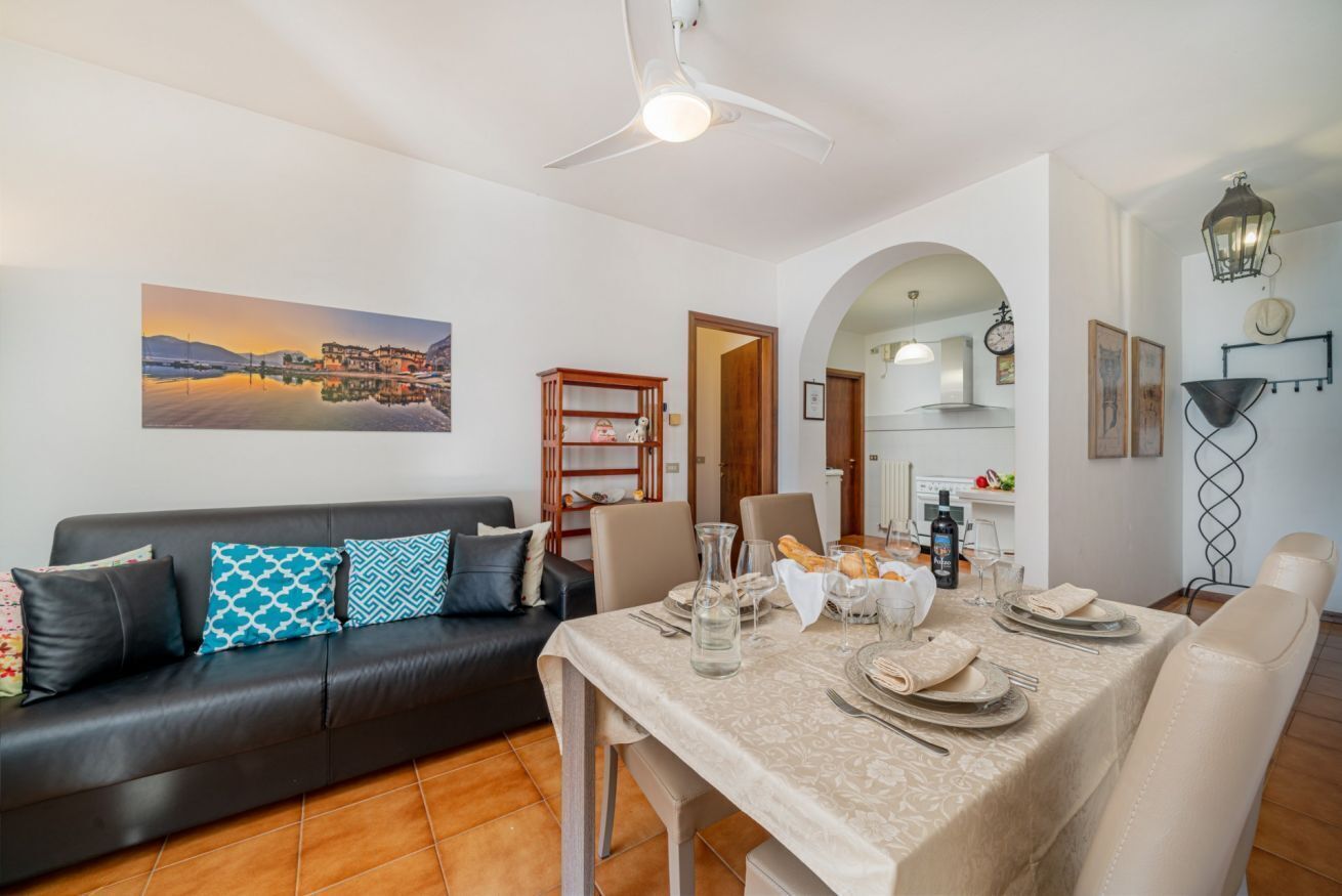 A dining area with a table set for two, a sofa with patterned cushions, a ceiling fan, and a cityscape painting on the wall.