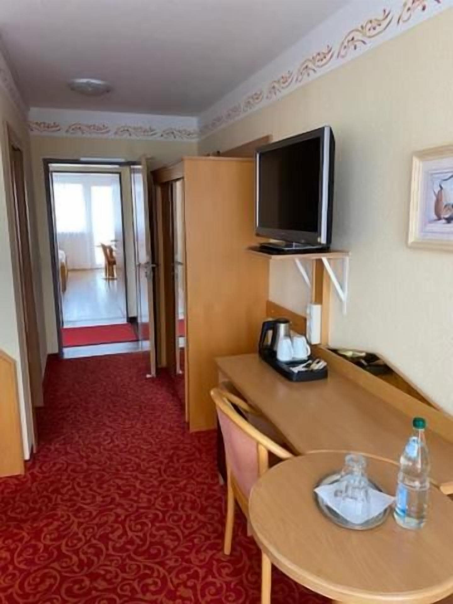 A hotel room with a television on a stand, a desk with a chair, a small round table, and a red carpeted floor.