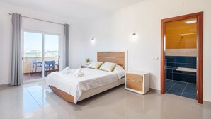 A modern hotel room with a bed, a bedside table, a small desk, a chair, and a sliding glass door to a balcony. - Casa Carmo - Villa with private pool, billiards and table tennis. (Carvoeiro)