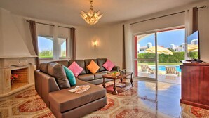 A spacious living room with a leather sofa, a fireplace, and a large window overlooking a pool area. - Villa in Alporchinhos (Porches) within walking distance to the beach. (Porches)