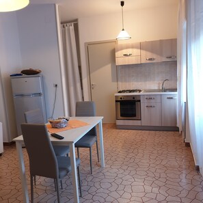 Una cocina compacta con estufa a gas, horno y lavabo. Hay una mesa de comedor con sillas y un refrigerador. El suelo presenta un diseño de baldosas geométricas.