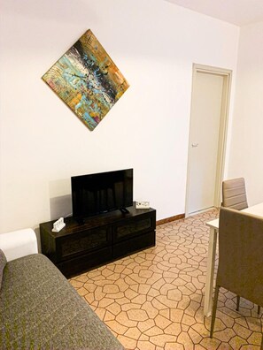 Un salón con televisor sobre un mueble de madera oscura, un cuadro abstracto y colorido en la pared, y suelo de baldosas.