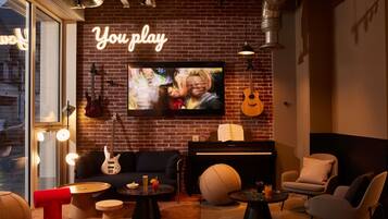 Ein gemütlicher Loungesessel mit Backsteinwand, einer Neonreklame, einem Fernseher, einer Gitarre, einem Klavier und bequemen Sitzgelegenheiten.