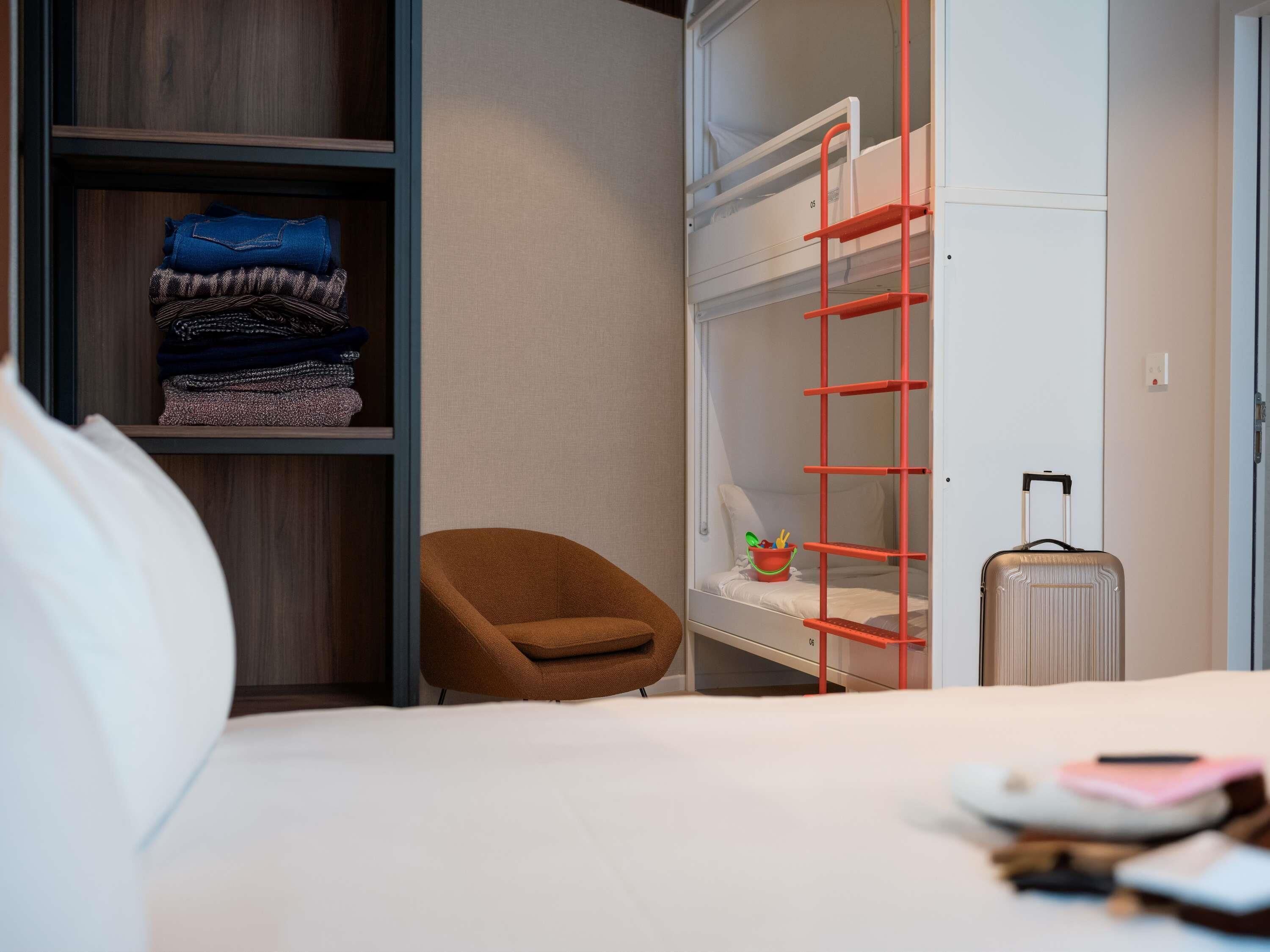 Habitación de hotel con literas, una silla marrón, una maleta y una cama con edredón blanco.