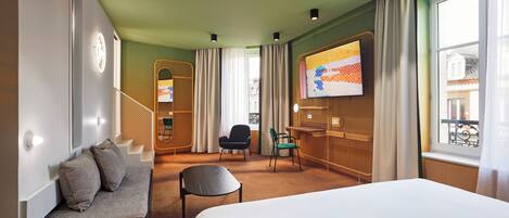 A hotel room with a bed, a seating area with cushions, a desk, a chair, a television, and a window with curtains.