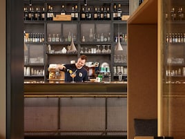 Zona de bar con estantes llenos de botellas, un barista preparando una bebida y una máquina de café.