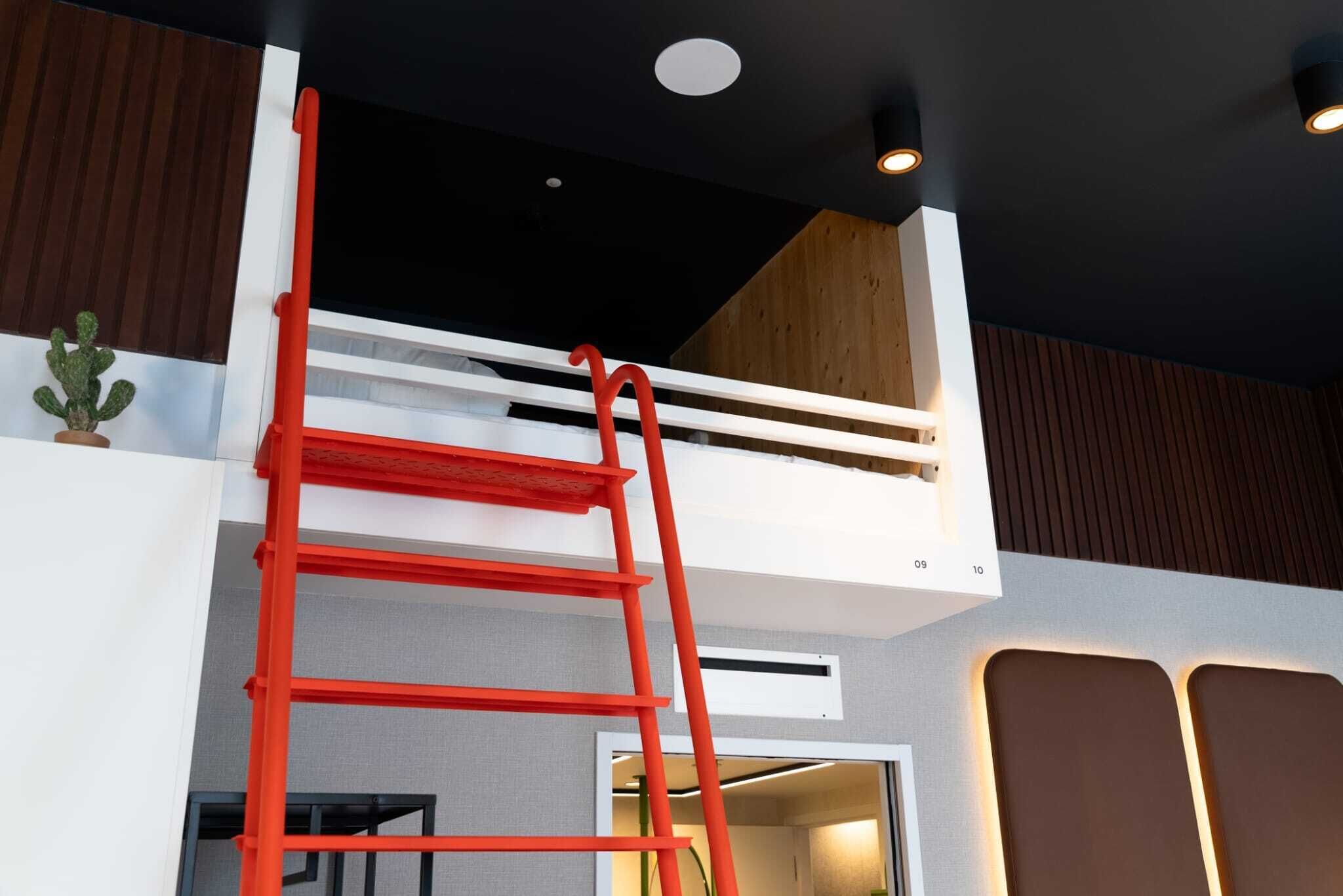 A red ladder leading to a mezzanine level with a white railing, a small potted plant, and recessed ceiling lighting.