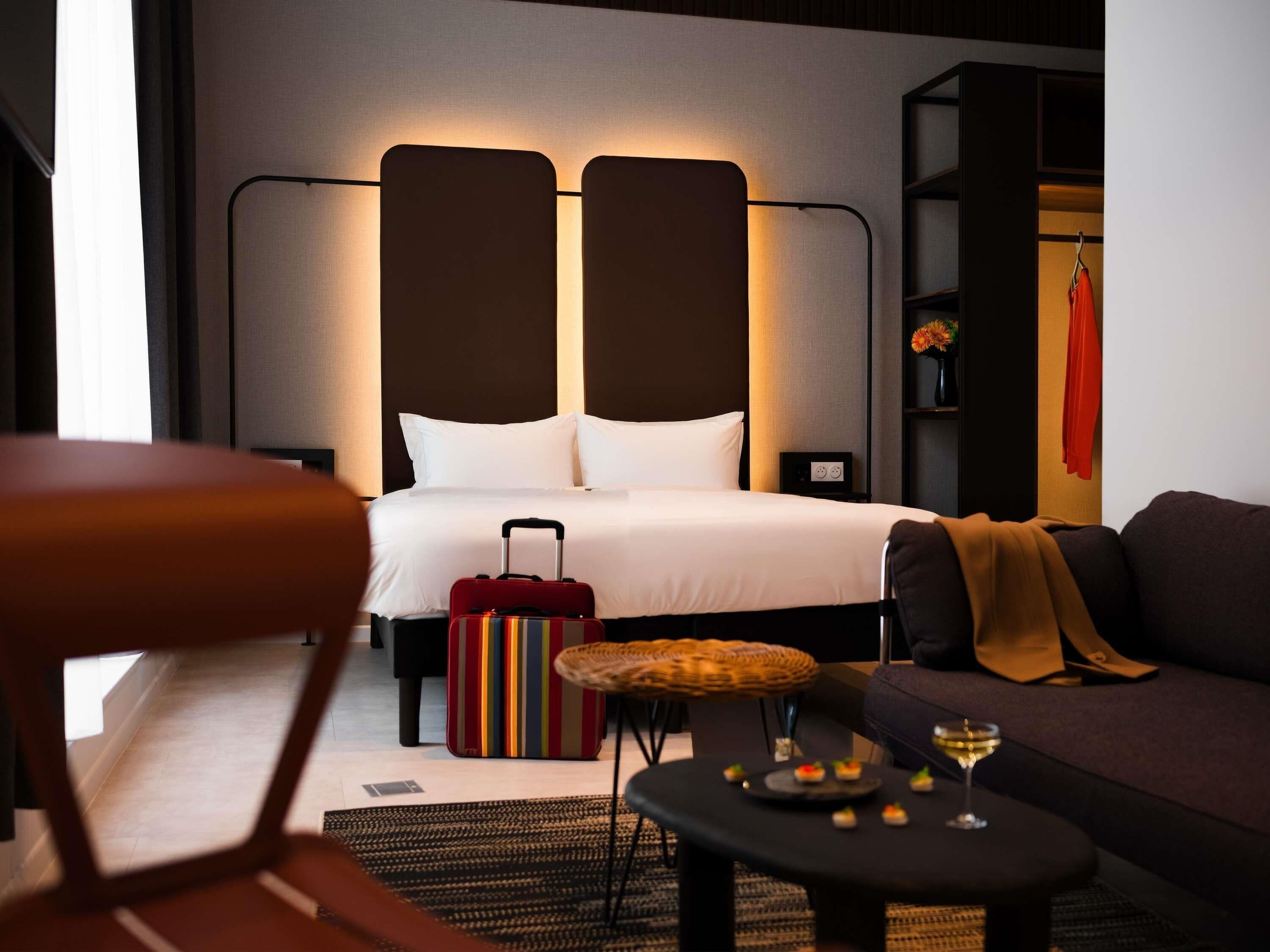 Habitación de hotel con cama, maleta, sofá, una mesita con un vaso y aperitivos, y una silla.