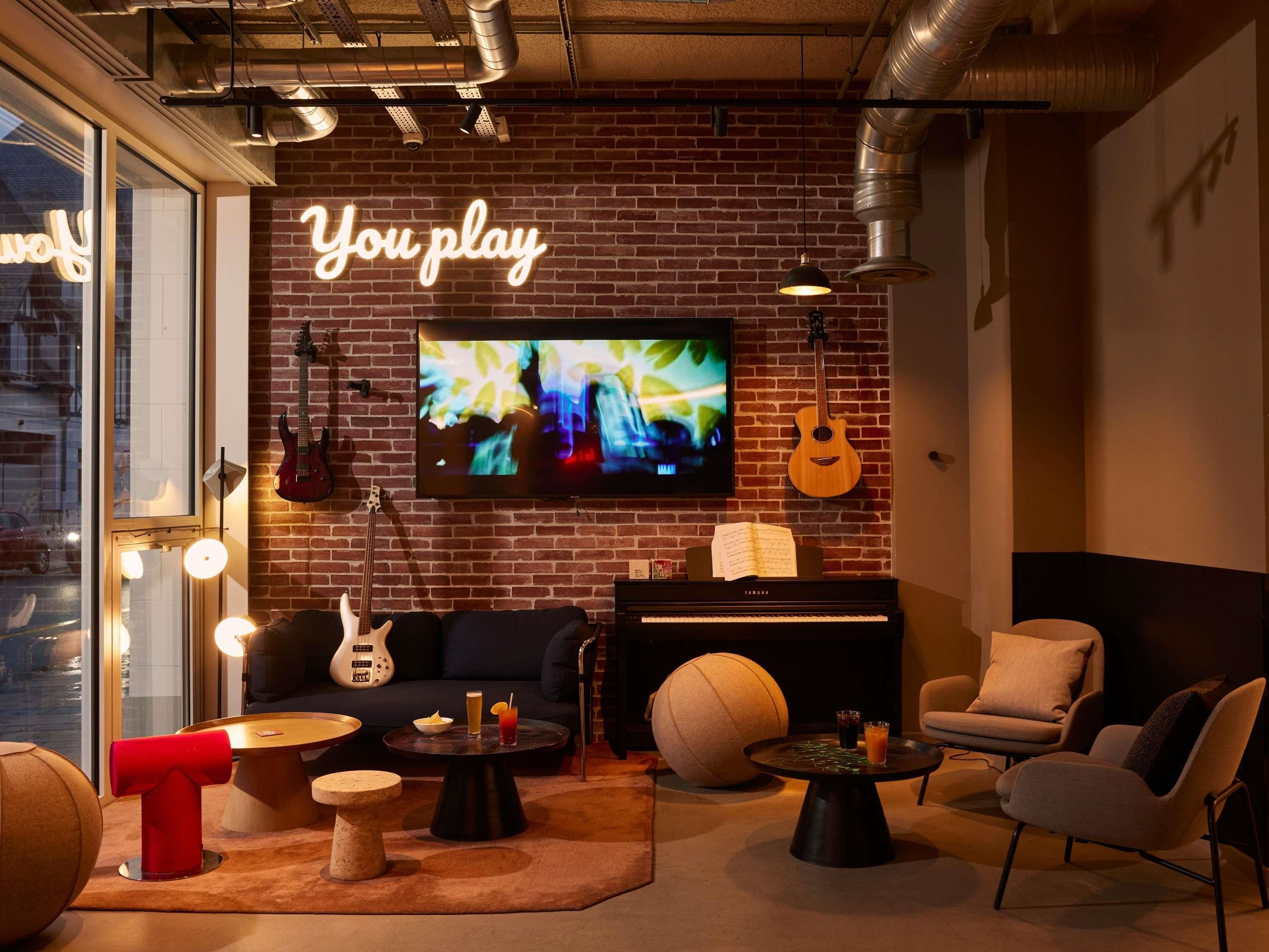 Un rincón acogedor con pared de ladrillo, televisión, guitarra, piano, asientos cómodos y un letrero de neón que dice 'You play'.