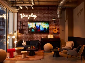Un rincón acogedor con pared de ladrillo, televisión, guitarra, piano, asientos cómodos y un letrero de neón que dice 'You play'.