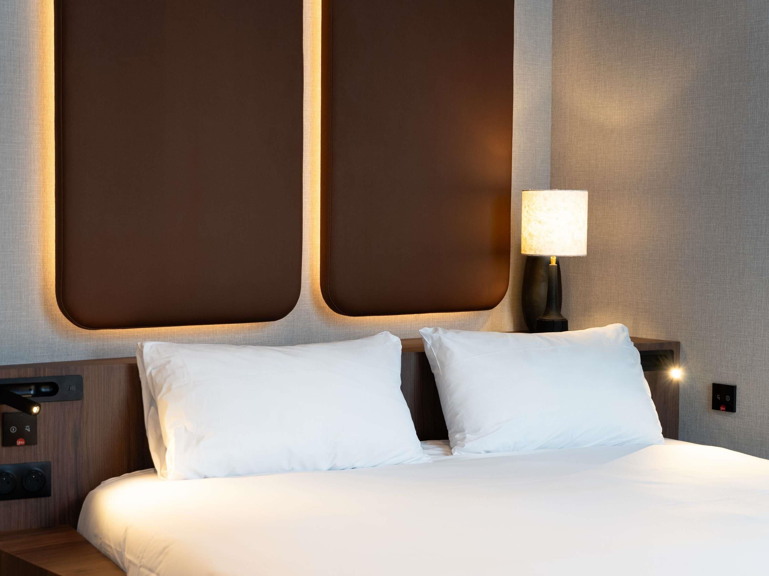 A hotel room with a large bed, two bedside tables, a wall-mounted lamp, and a control panel on the wall.