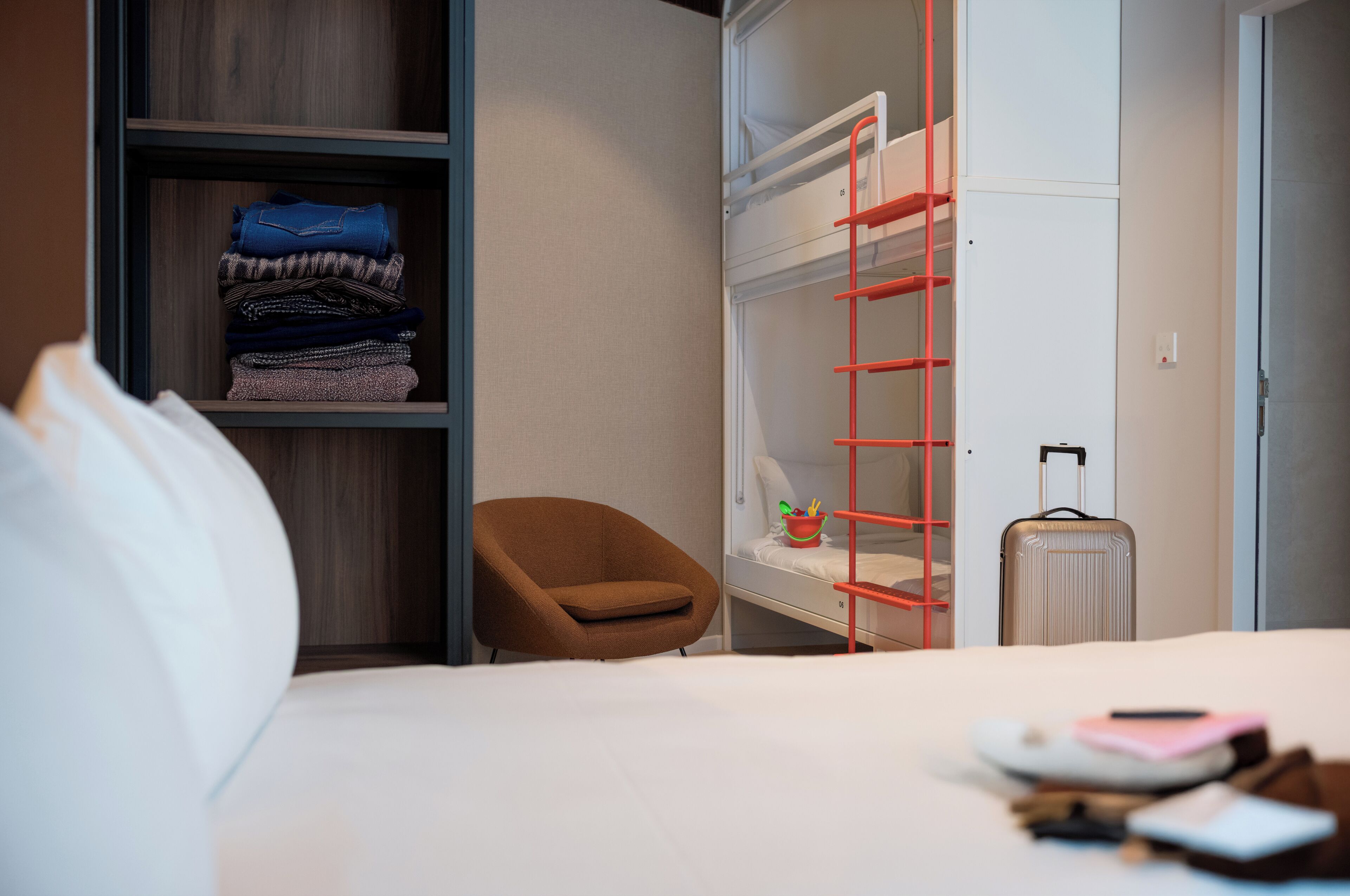 A hotel room with a bed, a brown chair, a wardrobe, a bunk bed, and a suitcase.