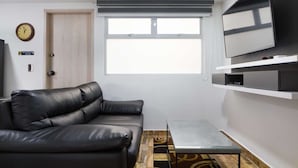 Een moderne woonkamer met een zwarte leren bank, een glazen salontafel en een tv aan een witte muur.