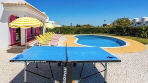 Una mesa de billar con superficie de fieltro azul y una paleta de tenis de mesa, rodeada de sillones de descanso y sombrillas de colores.