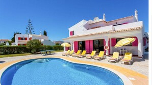 Una piscina con tumbonas y sombrillas, rodeada por un edificio blanco con contraventanas rosas.