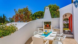 Un espace de restauration en plein air avec une table blanche dressée pour quatre personnes, entourée de chaises blanches, d’un barbecue intégré et d’un foyer.