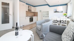 A modern kitchen with white cabinets and a built-in oven, a dining area with a white table and chairs, and a grey sofa in the living area. - Cozy apartment with balcony for 4 people (Greetsiel)