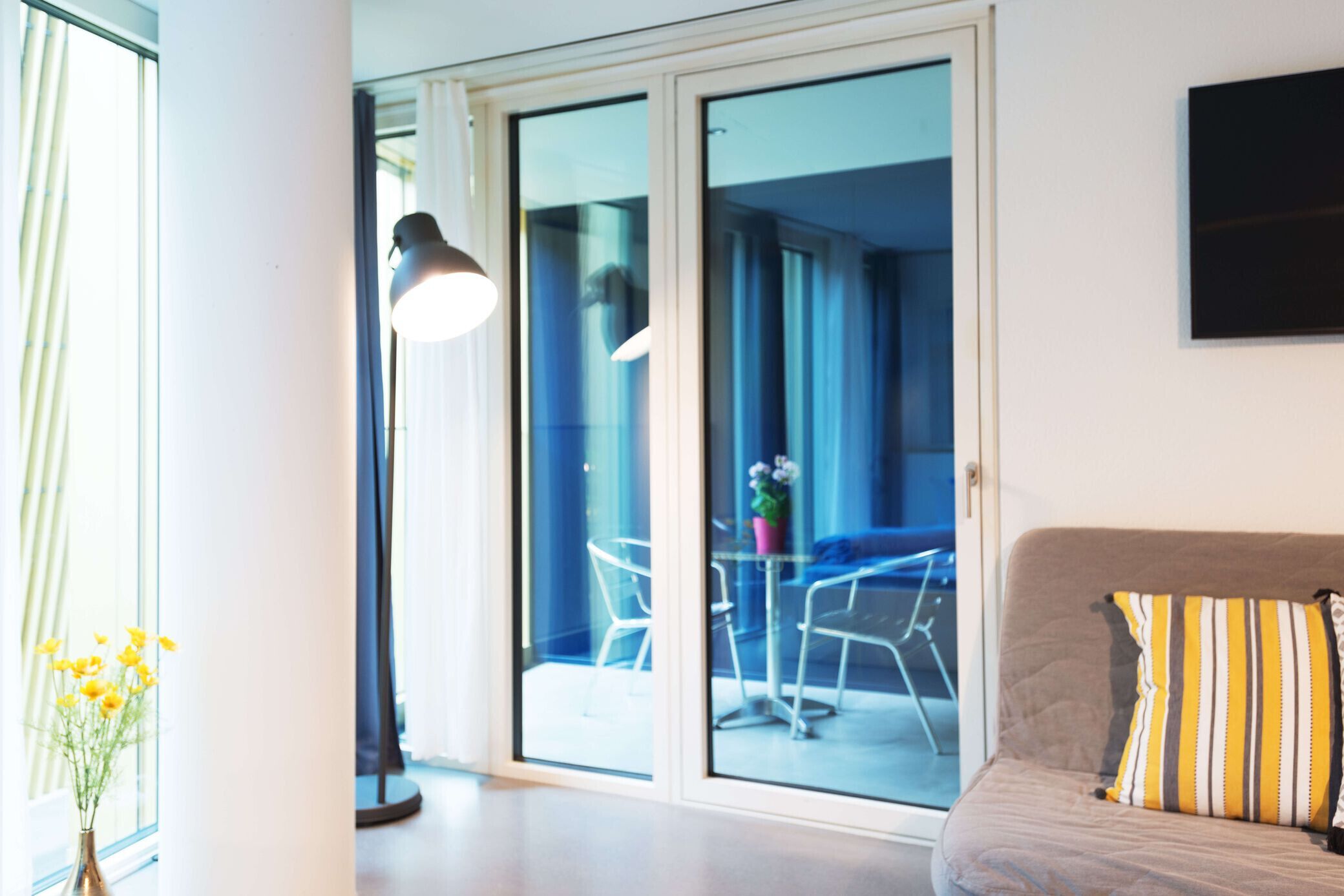 A modern living room with a sofa, a lamp, and a view through a glass door to an outdoor seating area.