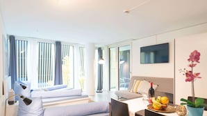 Habitació d'hotel moderna amb un llit sofà, una taula de dinar amb cadires, una televisió i una finestra gran amb cortines.