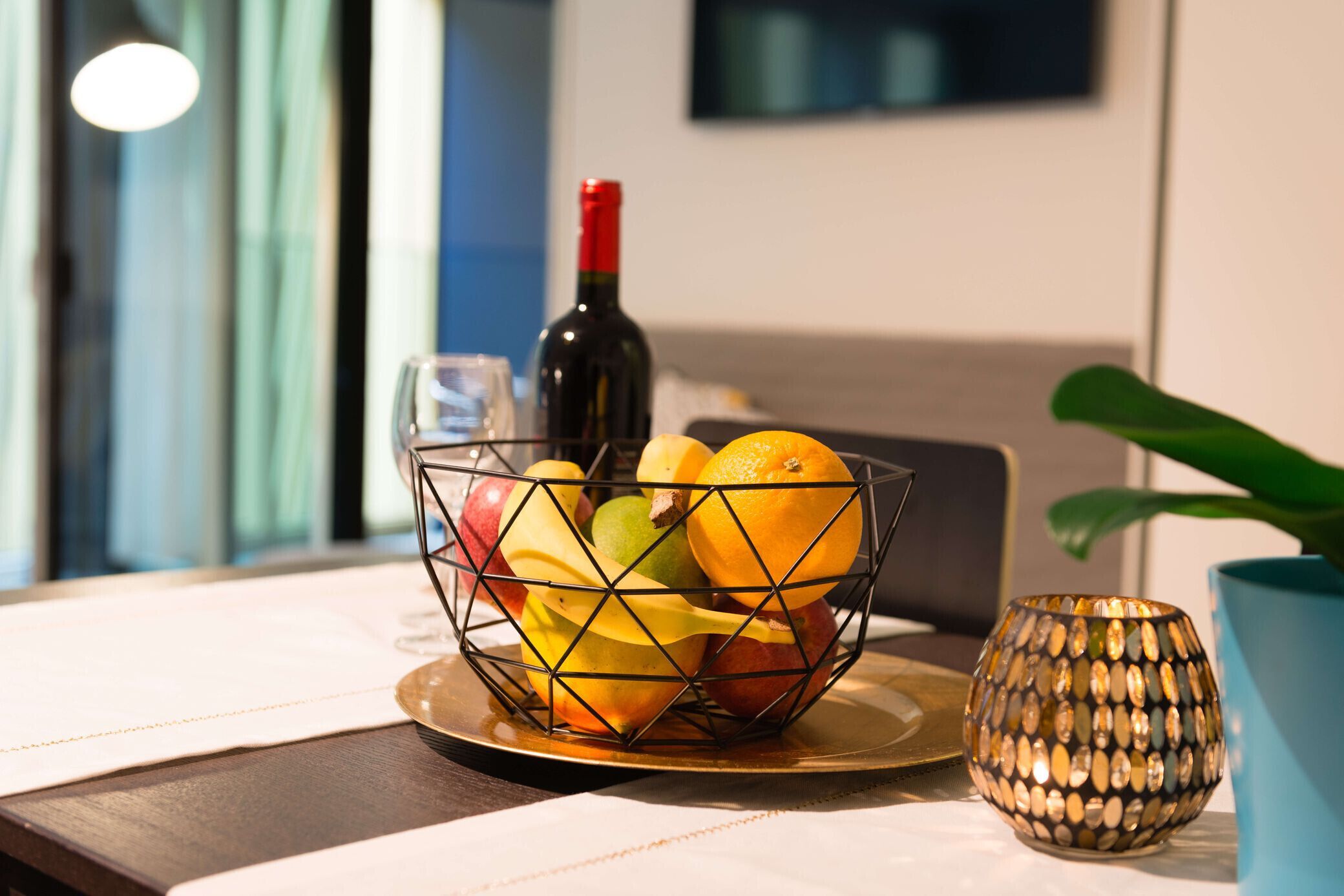 Un panier de fruits est posé sur une table, à côté d’une bouteille de vin et d’une bougie décorative.