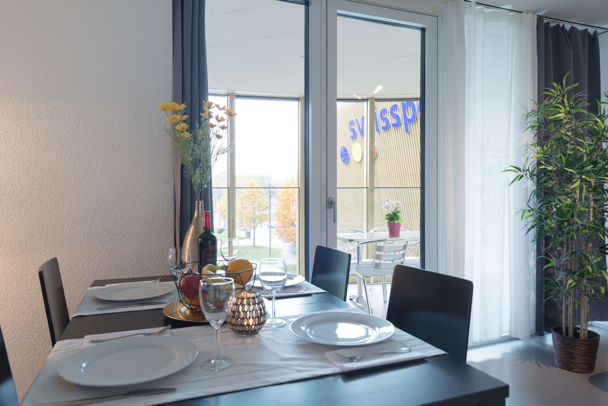 A dining area with a table set for two, a potted plant, and a view of the outside.