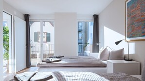 Un dormitorio moderno con una cama grande, una mesita de noche con lámpara y vistas a un balcón con un edificio al fondo.