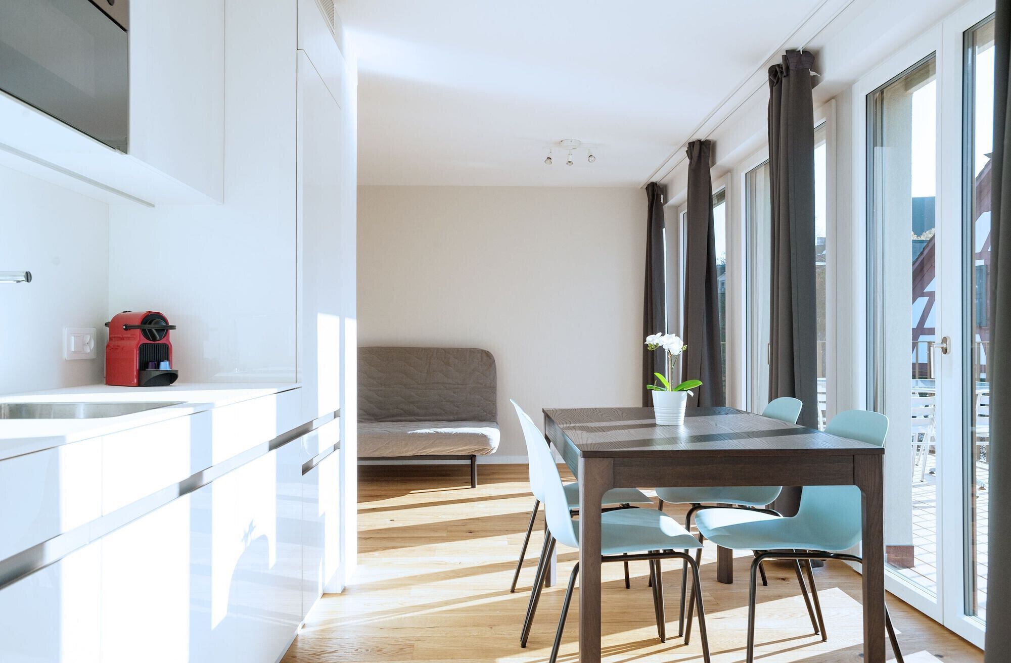 Una cocina moderna con horno empotrado, una zona de comedor con mesa y sillas de madera, y ventanas grandes con cortinas.
