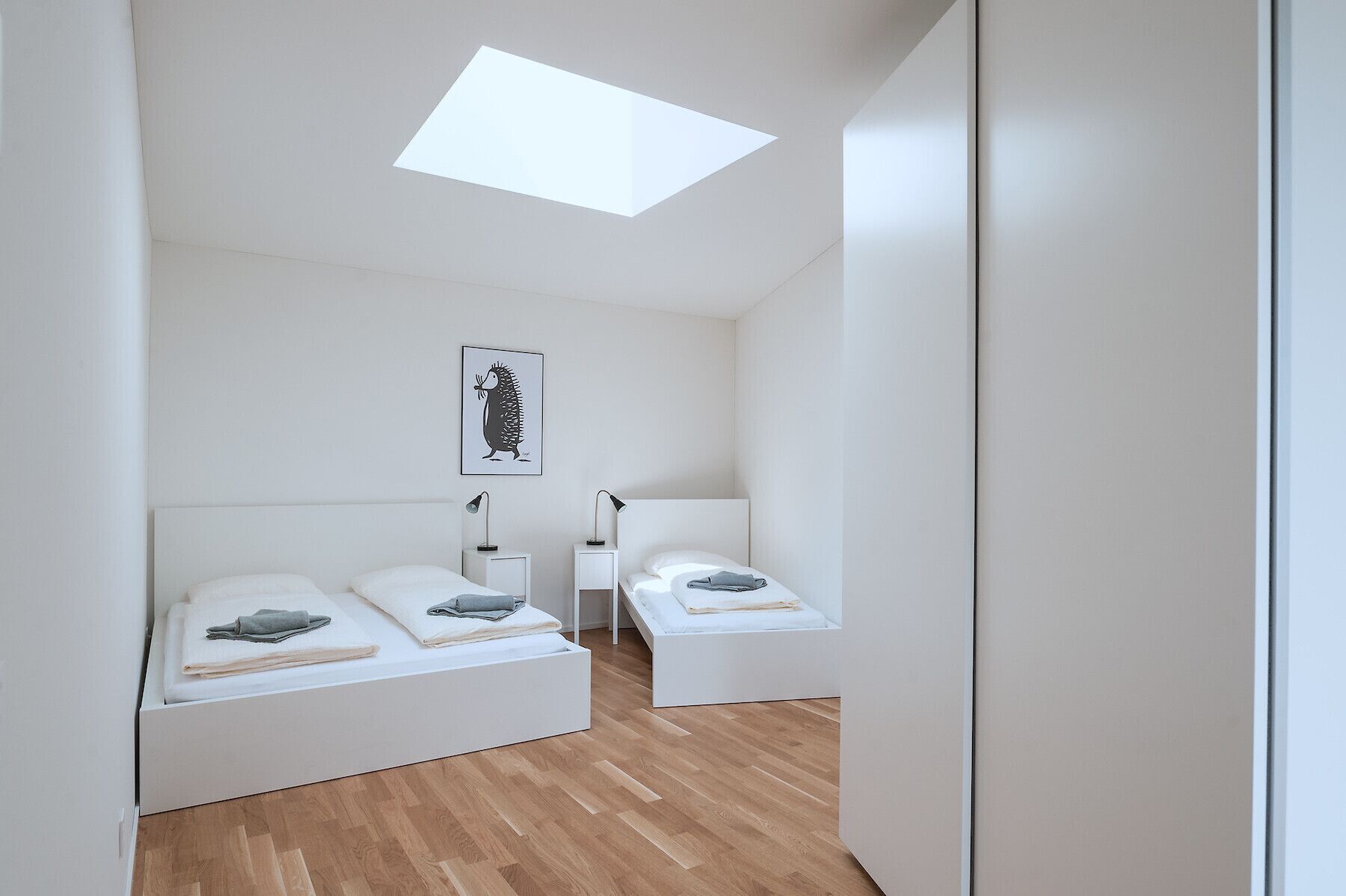 Ein modernes, minimalistisches Schlafzimmer mit zwei Betten, einem Oberlicht und einem gerahmten Kunstwerk an der Wand.
