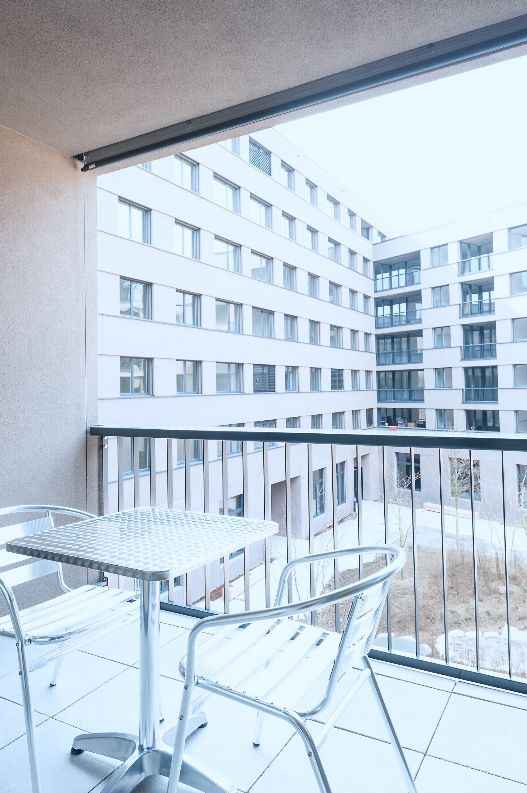 A balcony with a metal table and chairs, overlooking a modern building with multiple windows.