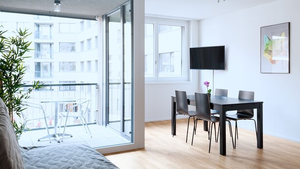 A modern living room with a balcony, a dining area with a table and chairs, a flat-screen TV mounted on the wall, and a large window providing a view of the cityscape. - BS Fox I - Messe Hitrental Apartment (Basel)