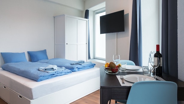 Una habitación de hotel con una cama, un escritorio con una silla, una mesa de comedor, un televisor montado en la pared y una ventana con cortinas.