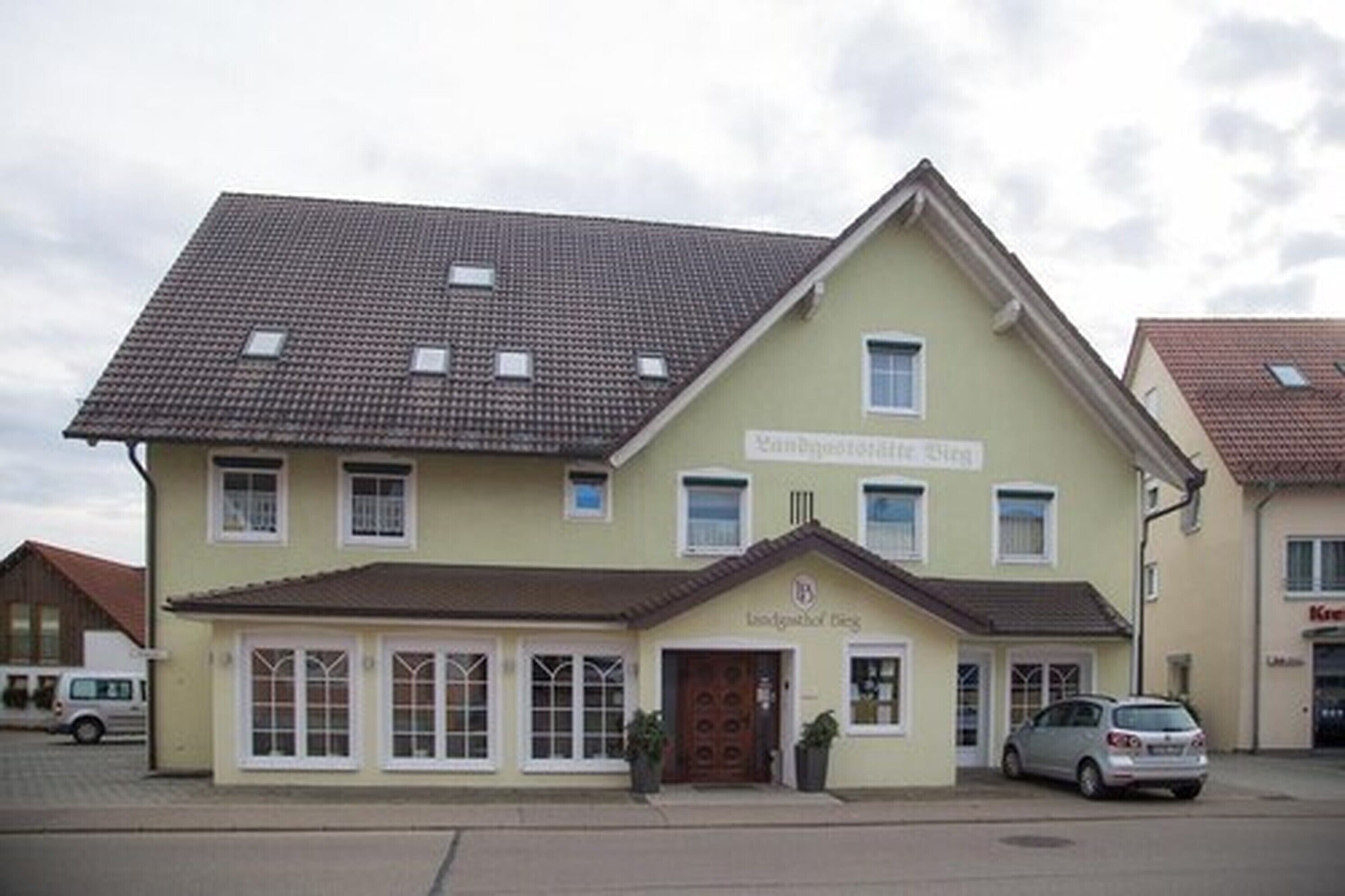 A two-story building with a tiled roof, a sign reading "Hotel Restaurant," and a parking area with cars.