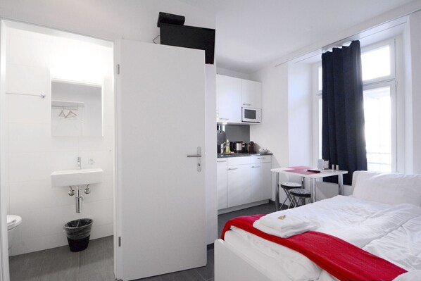 A hotel room with a bed, a small kitchenette, a bathroom, and a window with curtains.