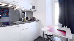 A compact kitchen with white cabinets, a microwave, a kettle, and a small table with a red mat. - LU Station II - Hitrental Apartment (Lucerne)