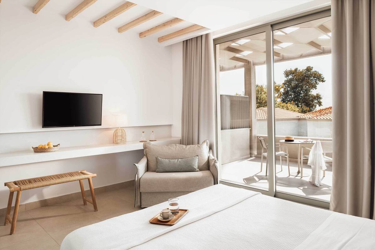 Une chambre d’hôtel moderne dotée d’un grand lit, d’un téléviseur à écran plat fixé au mur, d’une petite table avec un plateau, et d’un balcon avec une table et des chaises.