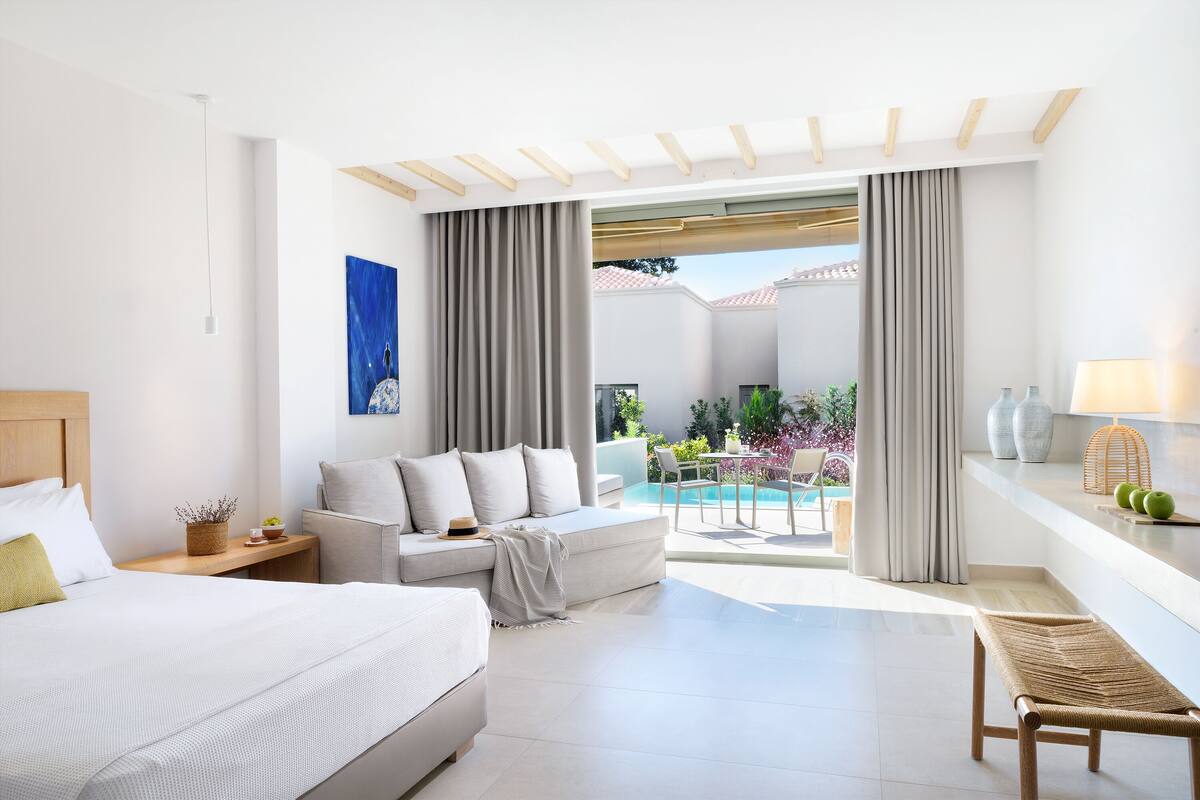 Une chambre d’hôtel moderne équipée d’un lit, d’un canapé, d’une banquette, d’une table à manger et offrant une vue sur une piscine et un jardin.