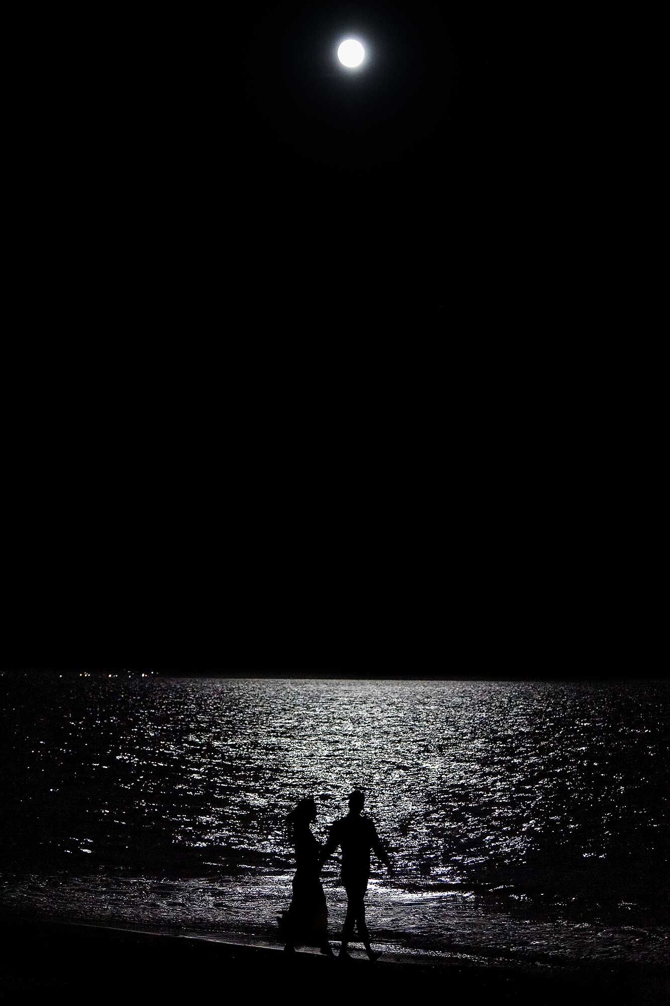 A night scene with a full moon and silhouettes of two people holding hands on a beach.