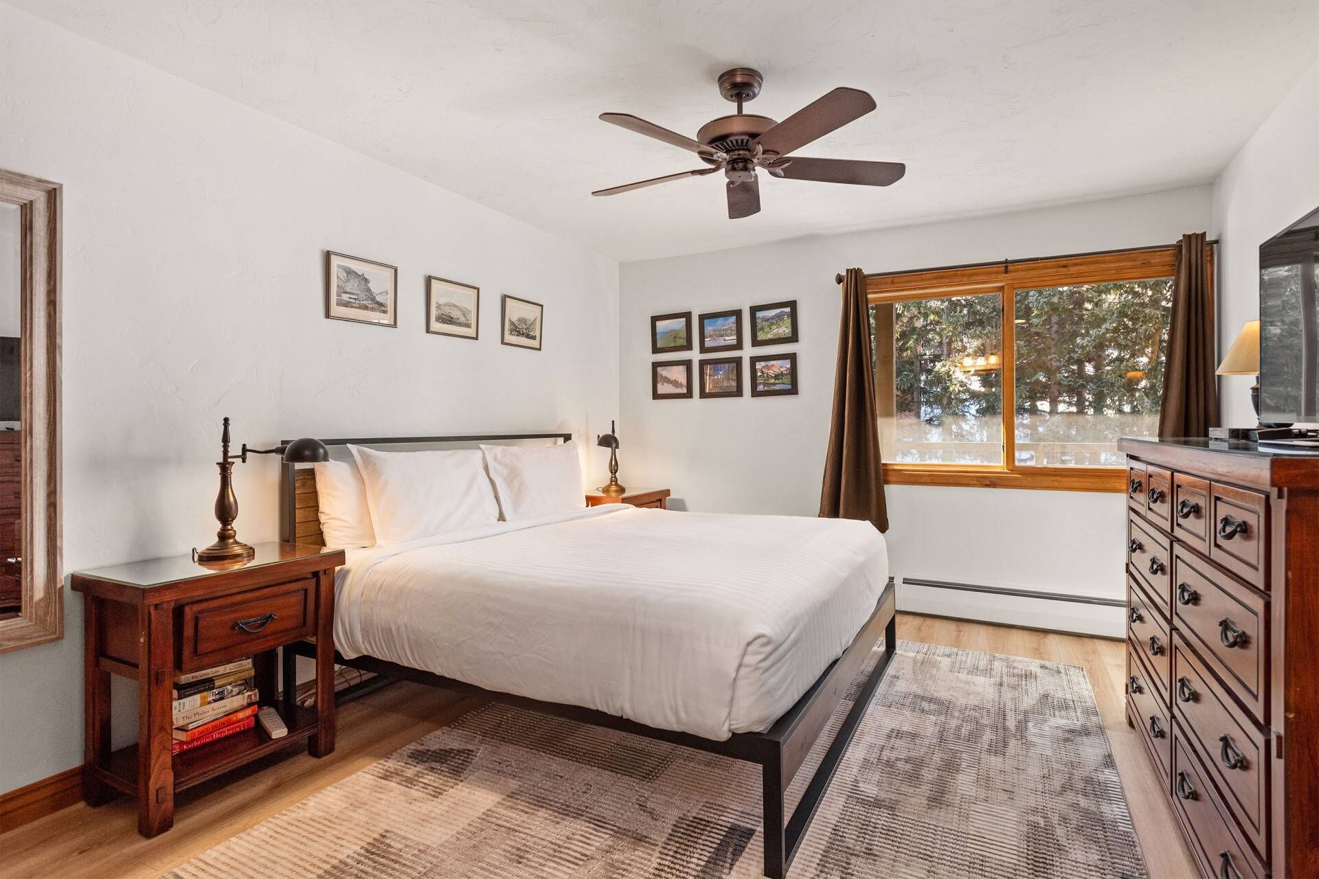 A bedroom with a bed, wooden nightstands, a dresser, a ceiling fan, framed pictures on the wall, and a window with curtains.