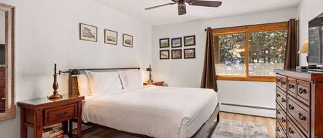 A bedroom with a bed, wooden nightstands, a dresser, a ceiling fan, framed pictures on the wall, and a window with curtains.