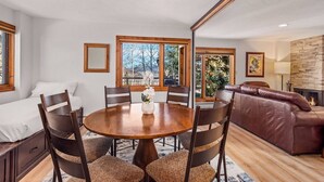 A dining area with a round wooden table, six chairs, and a vase of flowers. There is a brown leather sofa and a fireplace in the background. - Summer Haven in Snowmass! Stunning Balcony Views, Pool/Hot Tub, Wood FP, Sleeping Den, W/D & Parking (Snowmass Village)